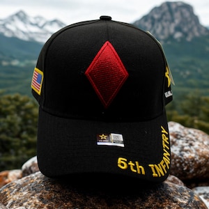 May include: Black baseball cap featuring a red diamond emblem and the words "5th INFANTRY" in gold lettering. The cap also has a small American flag patch and a U.S. Army patch. The cap is set against a mountain backdrop.