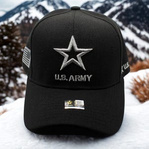 Op de afbeelding: Zwarte baseball cap met een wit geborduurde ster en de tekst "U.S. ARMY". De cap heeft een gebogen klep en een kleine Amerikaanse vlag patch. Een klein label is zichtbaar op de klep. De achtergrond toont een besneeuwde berg.