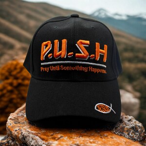 May include: Black baseball cap featuring the embroidered text "P.U.S.H. Pray Until Something Happens" in orange. A small fish symbol is embroidered on the side. The cap is set against a blurred outdoor background.