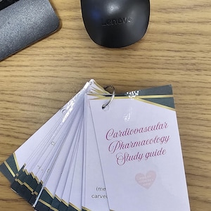 May include: A set of flashcards with the title "Cardiovascular Pharmacology Study guide" in elegant script. The cards are white with gold and dark green accents. A black Lenovo mouse and part of a keyboard are in the background.