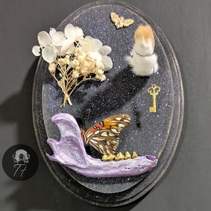 May include: An oval-shaped, dark background with a purple jawbone, a butterfly, dried flowers, a small furry creature, a gold key, and a gold butterfly. The jawbone has gold teeth and the butterfly has orange and brown wings. The background has a speckled, starry effect.