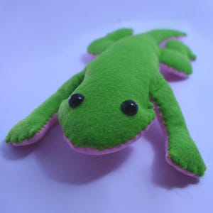 May include: A plush green frog toy with black button eyes and pink accents on the feet and underside. The frog is lying on a light purple surface, with its arms and legs extended. The toy is made of a soft, fuzzy material.