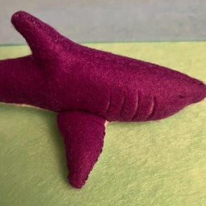 May include: A handmade felt shark in a deep purple color. The shark has a dorsal fin, a pectoral fin, and a tail fin. The shark's body has stitched gill slits. The shark is resting on a green surface.
