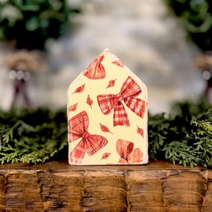 May include: A house-shaped wooden block with a cream-colored background featuring a repeating pattern of red plaid bows and small scattered leaves. The object has a pointed roofline and a flat base, displaying a decorative print on the front surface.