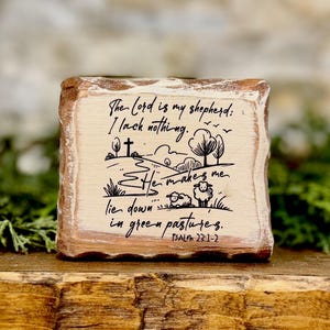 May include: A rustic wooden block with a light-colored surface and a brown, textured edge. The block features a black illustration of a landscape with sheep and trees, and the text "The Lord is my shepherd: I lack nothing." Psalm 23:1-2 is also written.