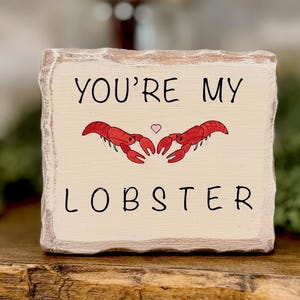 May include: A cream-colored wooden block with the text "YOU'RE MY LOBSTER" in black. Two red lobster illustrations face each other above the word "LOBSTER", with a small pink heart between them. The block has a rustic, distressed look.