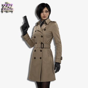 Op de afbeelding: Een beige dubbelrijïge trenchcoat met een bijpassende riem, donkere knopen en epauletten. De jas heeft een klassieke kraag, lange mouwen met gespen aan de manchetten en een getailleerde pasvorm. Het ZAQ Studios logo staat linksboven in beeld.