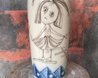 Könnte beinhalten: Eine weiße Keramikvase mit einer skurrilen Illustration eines Mädchens in einem Kleid. Die Vase hat blaue geometrische Muster oben und unten. Das Mädchen ist in Schwarz und Grau gezeichnet, mit einem traurigen Ausdruck. Die Vase steht auf einem Metallständer.