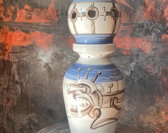 Könnte beinhalten: Eine hohe, handgefertigte Keramikvase mit einem einzigartigen, gestapelten Design. Die Vase hat ein weißes, blaues und braunes Farbschema mit einer skurrilen Illustration auf dem unteren Teil. Die Vase steht auf einer Metalloberfläche.