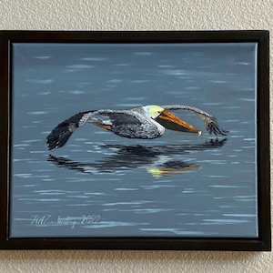 May include: A framed painting depicts a pelican in flight over water. The bird's wings are outstretched, and its reflection is visible in the water. The pelican has a yellow head and a long, orange beak. The artwork is signed and dated 2022.