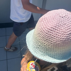 Op de afbeelding: Een gehaakte bucket hat met een verloop van roze, beige en lichtblauw. Een persoon draagt de hoed en houdt een gele beker met een rietje vast. Een andere persoon in een wit t-shirt en donkere shorts is op de achtergrond.