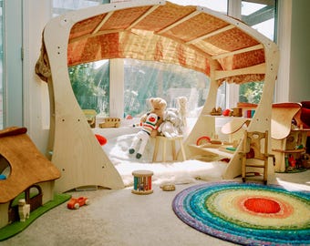 May include: A wooden playhouse with a canopy and shelves, filled with toys and stuffed animals. A colorful rug and miniature wooden houses are in the foreground. The playhouse has a light wood frame and a patterned fabric canopy.