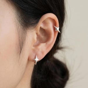 Puede incluir: Primer plano de una oreja adornada con dos pendientes plateados en forma de estrella. Uno es un pendiente de botón y el otro es un brazalete que se envuelve alrededor de la parte superior de la oreja. La joyería tiene un diseño minimalista y un acabado brillante y reflectante.