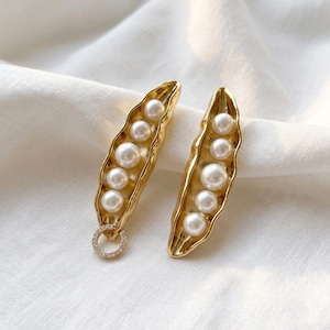 May include: A pair of gold-colored earrings, each shaped like a pea pod, with six white pearl beads inside. One earring has a small, round, diamond-like accent at the bottom. The earrings are displayed on a white, textured surface.