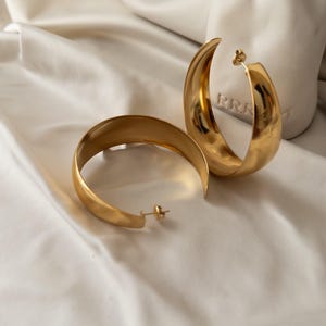 Peut inclure: Une paire de boucles d'oreilles créoles dorées. Les boucles d'oreilles ont un design lisse et incurvé et une fermeture à tige. Elles sont présentées sur une surface blanche et texturée.