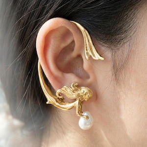 May include: Gold-toned ear cuff featuring a mermaid design. The cuff wraps around the ear and includes a mermaid figure holding a white pearl. The design has a flowing, organic shape.