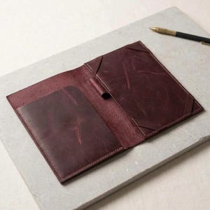 May include: An open, dark burgundy leather passport holder featuring a smooth, matte finish with visible natural grain textures. The interior includes a vertical pocket on the left side and two elastic corner straps on the right side for securing documents.