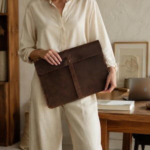 May include: A dark brown leather laptop sleeve featuring a rectangular shape with rounded corners and a vertical strap closure that secures the flap. The surface has a matte, distressed texture with visible natural grain and stitching details.