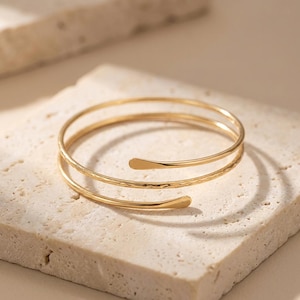 May include: A gold arm cuff bracelet with a layered design. The bracelet has two smooth bands and one textured band. The ends of the bracelet have a flattened, curved shape. The jewellery is displayed on a light-coloured, textured surface.