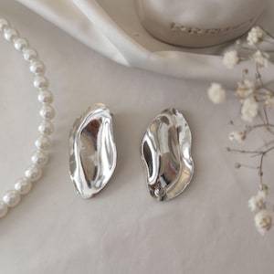 Może przedstawiać: Para srebrnych kolczyków w kształcie abstrakcyjnym, w kolorze srebrnym. Kolczyki mają falisty, organiczny kształt i błyszczącą, odbijającą światło powierzchnię. Szereg białych pereł jest widoczny po lewej stronie, a białe kwiaty po prawej.