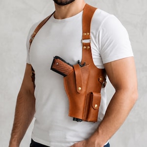 Könnte beinhalten: Ein hellbraunes Schulterholster aus Leder mit einem Hauptfach für eine Schusswaffe mit Holzgriff, einer Zusatztasche für ein Magazin, verstellbaren Riemen und goldfarbenen Metallbeschlägen, getragen über einem schlichten weißen T-Shirt.