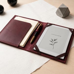 Puede incluir: Una funda de cuero rojo oscuro que contiene una tableta digital Supernote con un gráfico botánico en la pantalla, una pila de papel color crema en un bolsillo lateral y un bolígrafo metálico en un lazo central, asegurada con bandas elásticas.