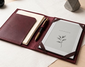 Pode incluir: Uma capa de couro vermelho escuro contendo um tablet digital Supernote com um gráfico botânico no ecrã, uma pilha de papel creme num bolso lateral e uma caneta metálica num suporte central, fixada por bandas elásticas nos cantos.