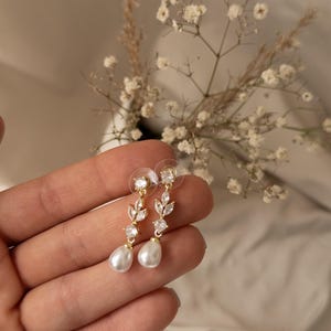 Puede incluir: Pendientes colgantes dorados con cristales transparentes y brillantes y perlas blancas en forma de pera. Los pendientes presentan un diseño en forma de hoja con cierre de poste. Las joyas se exhiben sobre un fondo suave y neutro.