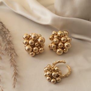 May include: Gold-colored jewelry set featuring a pair of cluster earrings and a matching ring. The earrings and ring are composed of numerous small, spherical beads clustered together. The jewelry is displayed on a cream-colored, satin-like fabric.