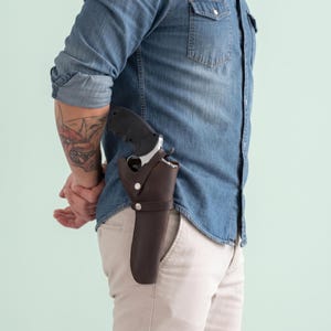 Può includere: Una fondina in pelle marrone scuro fissata con due bottoni a pressione in metallo argentato, contenente un revolver nero e argento. La fondina è indossata sul fianco sopra una camicia di jeans blu abbinata a pantaloni chiari.