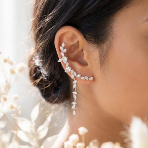 May include: A silver ear cuff featuring a delicate leaf design. The ear cuff curves along the ear and dangles down with a series of small, sparkling stones. The jewelry is set against a soft, blurred background.