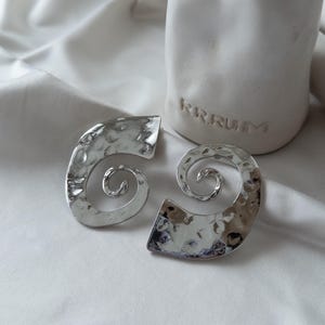 May include: A pair of silver-toned, hammered-metal earrings in a spiral design. The earrings have a textured surface and a polished finish, reflecting light. The earrings are displayed on a white, silky fabric.