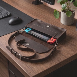 May include: A dark brown leather carrying case with a magnetic snap closure, containing a handheld gaming console with a black screen and blue and red controllers. The case features an adjustable leather shoulder strap with metal hardware.