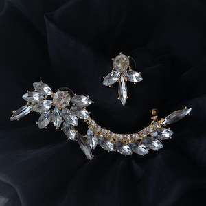 Può includere: Un set di due orecchini di cristallo su sfondo nero. Uno è un orecchino a lobo con un cristallo centrale e quattro accenti di cristallo. L'altro è un ear cuff dorato con più cristalli trasparenti.