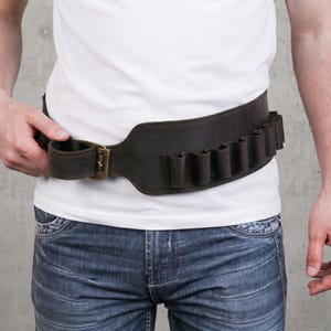 May include: A wide dark brown leather belt featuring a series of integrated loops along the side for holding small cylindrical items. The belt is secured with a brass-toned metal buckle and is worn over a plain white t-shirt and blue jeans.