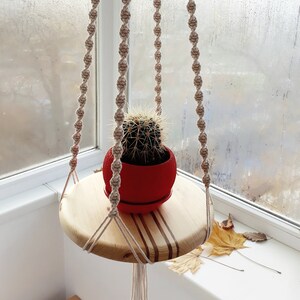 Può includere: Un portavasi in macramè con uno scaffale in legno e un vaso rosso con una pianta di cactus. Il portavasi è realizzato in corda di cotone naturale e ha una perlina di legno nella parte superiore.