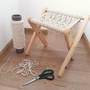 Op de afbeelding: Een houten kruk met een macram&eacute; zitting, een rol macram&eacute; koord en een schaar op een houten vloer.