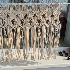 May include: A beige macrame wall hanging with a geometric pattern. The hanging is made of natural cotton cord and has a fringe at the bottom.