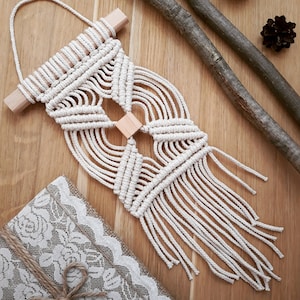 Könnte beinhalten: Ein Wandbehang aus Makramee mit geometrischem Muster, aus weißem Baumwollgarn und Holzleisten. Der Behang hat unten eine Fransenborte.