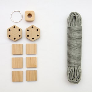 Puede incluir: Un conjunto de materiales de artesanía de madera para hacer un macetero de macramé. El conjunto incluye un anillo de madera, dos piezas hexagonales de madera con agujeros, seis piezas cuadradas de madera y un rollo de cuerda gris.