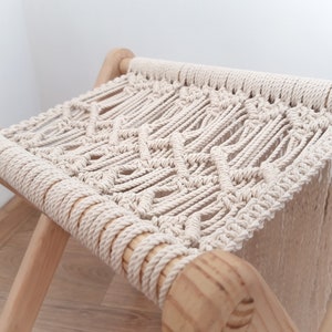 Op de afbeelding: Een witte macram&eacute; kruk met een houten frame. De kruk heeft een ruitpatroon en franjes aan de onderkant.