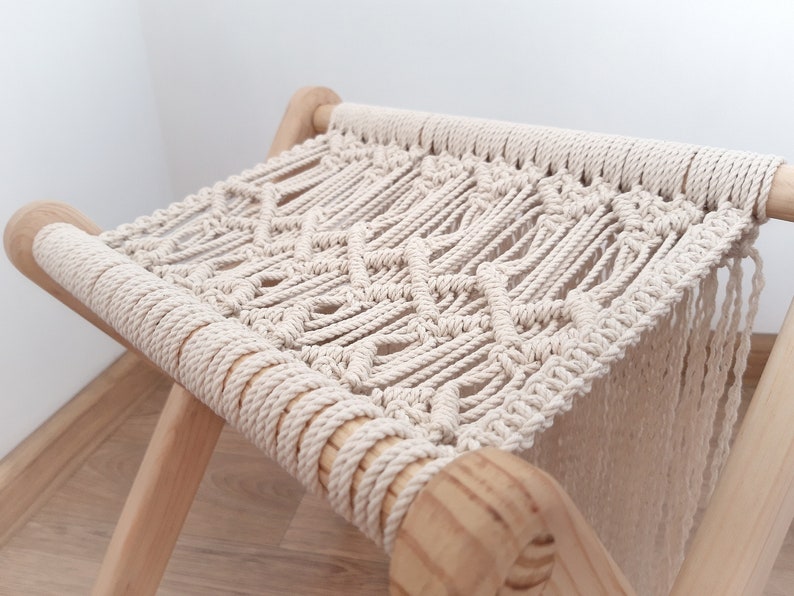 Op de afbeelding: Een witte macram&eacute; kruk met een houten frame. De kruk heeft een ruitpatroon en franjes aan de onderkant.