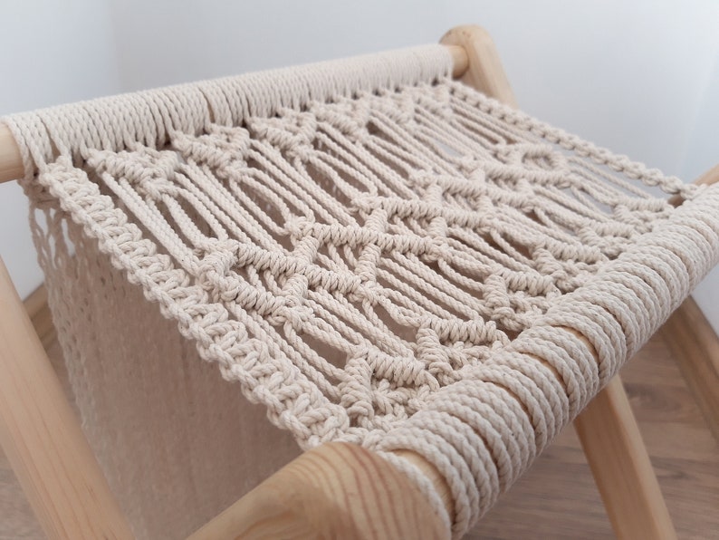 Op de afbeelding: Een macram&eacute; kruk met een zitting van natuurlijke beige touw en houten poten. De zitting is geweven in een ruitpatroon.