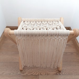 Op de afbeelding: Een macram&eacute; kruk met een bovenkant van natuurlijke witte katoenen koord en franjes. De kruk heeft een houten frame met een natuurlijke houtkleur.