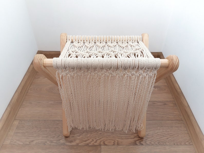 Op de afbeelding: Een macram&eacute; kruk met een bovenkant van natuurlijke witte katoenen koord en franjes. De kruk heeft een houten frame met een natuurlijke houtkleur.