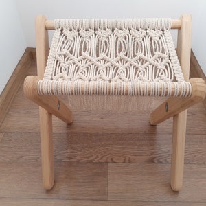 Op de afbeelding: Een kleine houten kruk met een macram&eacute; zitting. De kruk is gemaakt van natuurlijk hout en heeft een eenvoudig, rustiek ontwerp.
