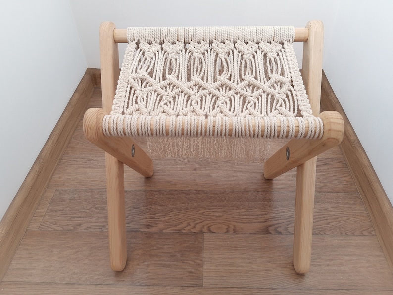 Op de afbeelding: Een kleine houten kruk met een macram&eacute; zitting. De kruk is gemaakt van natuurlijk hout en heeft een eenvoudig, rustiek ontwerp.