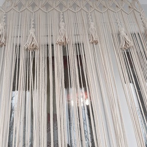 Puede incluir: Una cortina de macram&eacute; blanca con borlas cuelga en una ventana. La cortina est&aacute; hecha de cuerdas anudadas y tiene un borde superior decorativo.