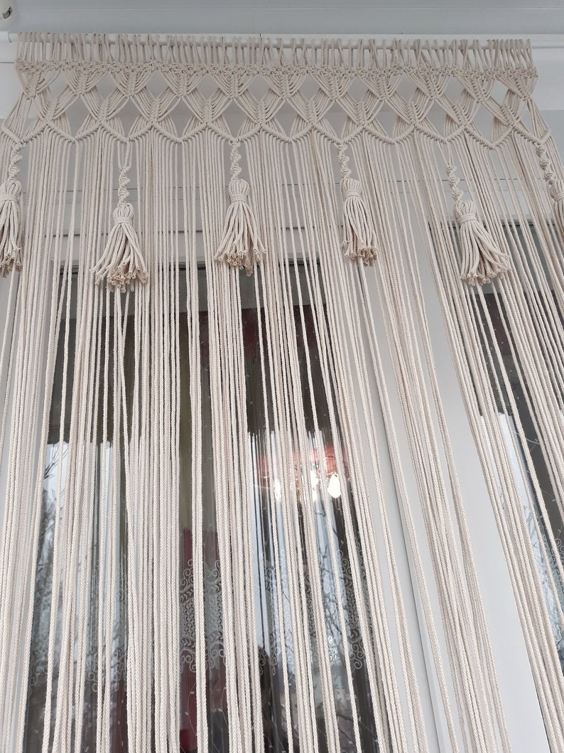 Puede incluir: Una cortina de macram&eacute; blanca con borlas cuelga en una ventana. La cortina est&aacute; hecha de cuerdas anudadas y tiene un borde superior decorativo.