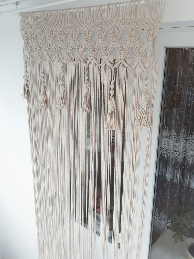 Puede incluir: Una cortina de macram&eacute; blanca con borlas que cuelgan en una ventana. La cortina est&aacute; hecha de cord&oacute;n de algod&oacute;n y tiene un patr&oacute;n geom&eacute;trico.